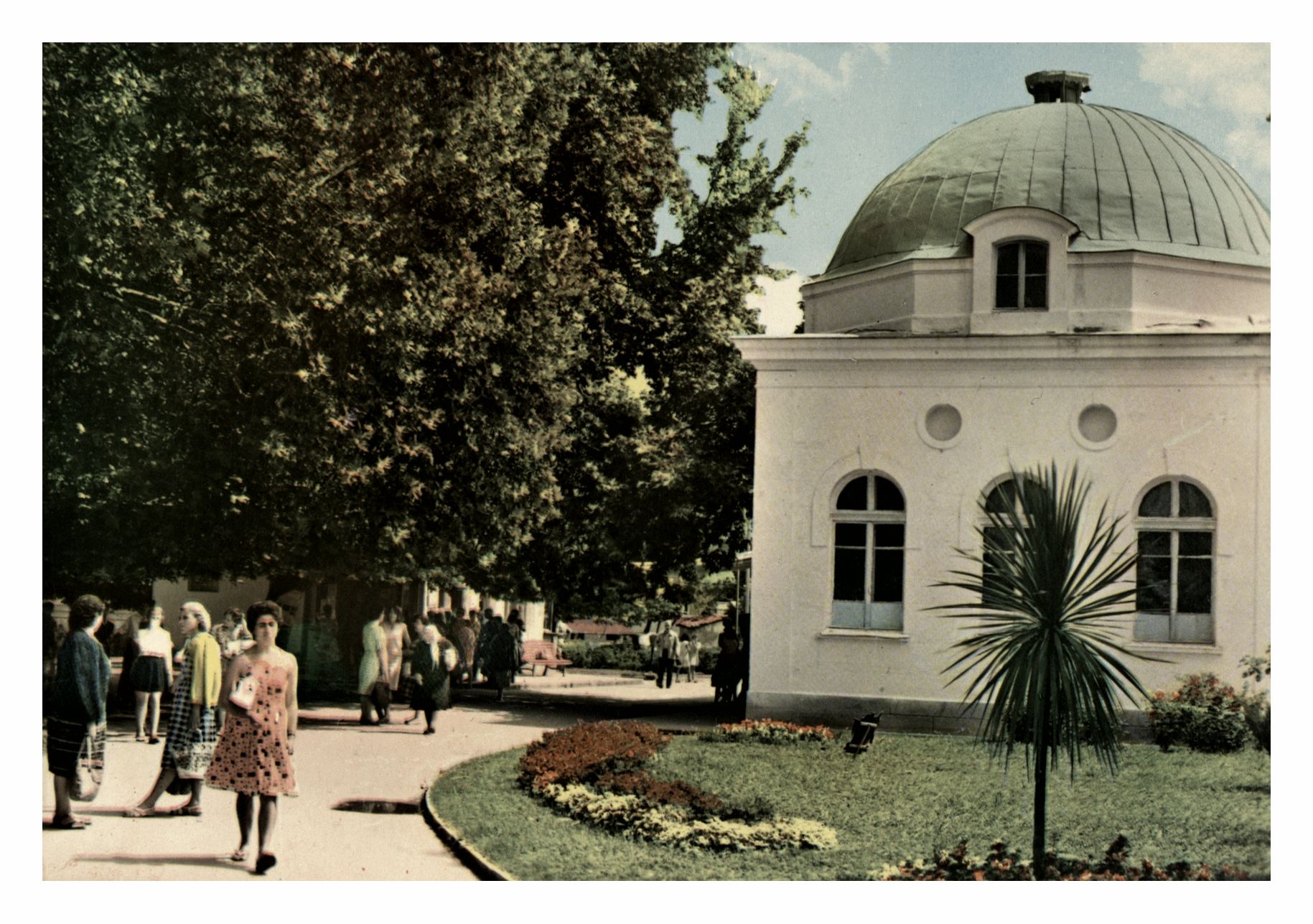 Архивна цветна фотография на градинката при Чифте баня в Кюстендил с алеи, цветни лехи, високи дървета и разхождащи се граждани. Вдясно се вижда сградата на Чифте баня с характерния ѝ купол.
