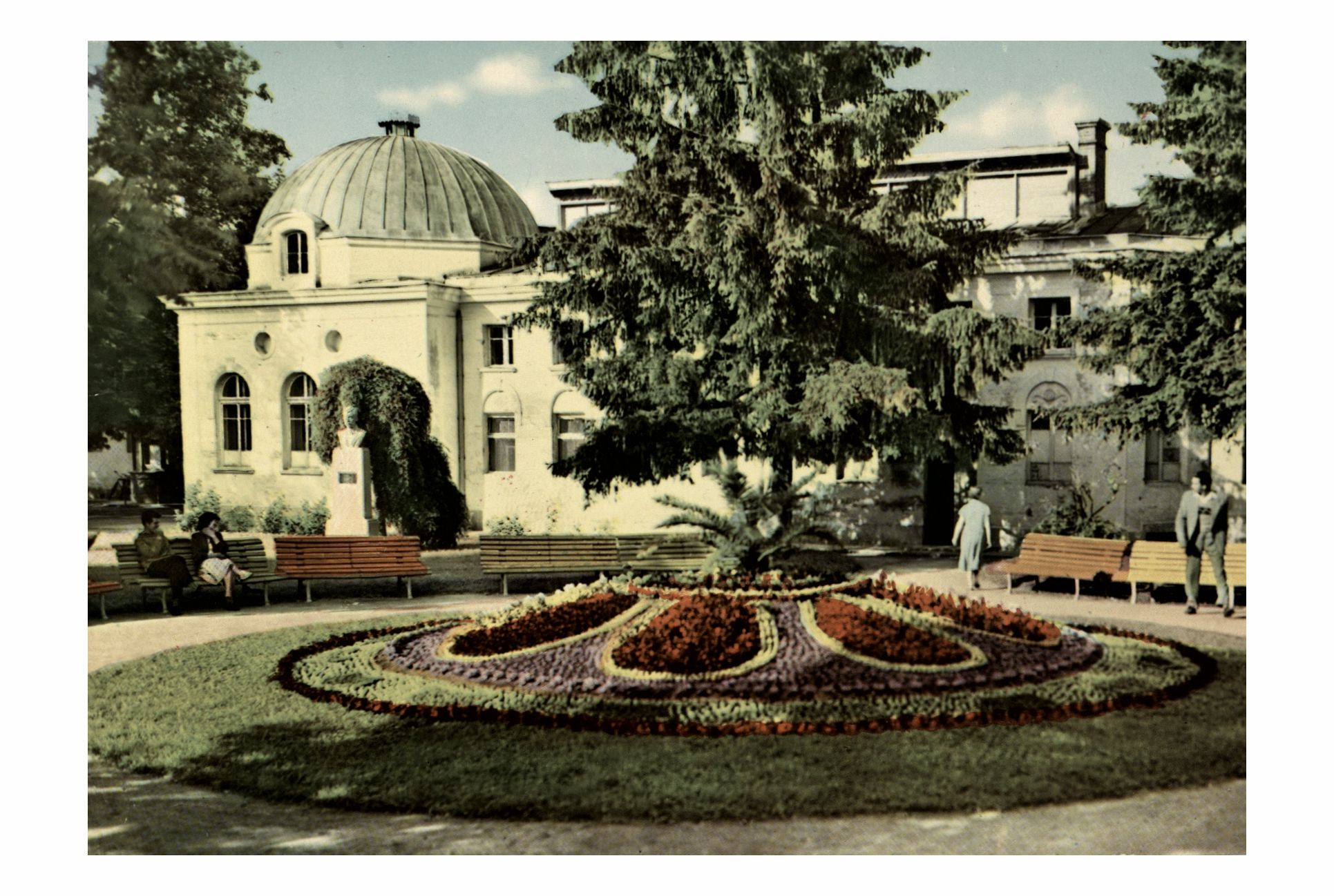 Архивна цветна фотография на градинката при Чифте баня в Кюстендил. На преден план се вижда голяма декоративна цветна леха с геометрични фигури, а на заден план – сградата на Чифте баня, пейки, посетители и високи дървета.